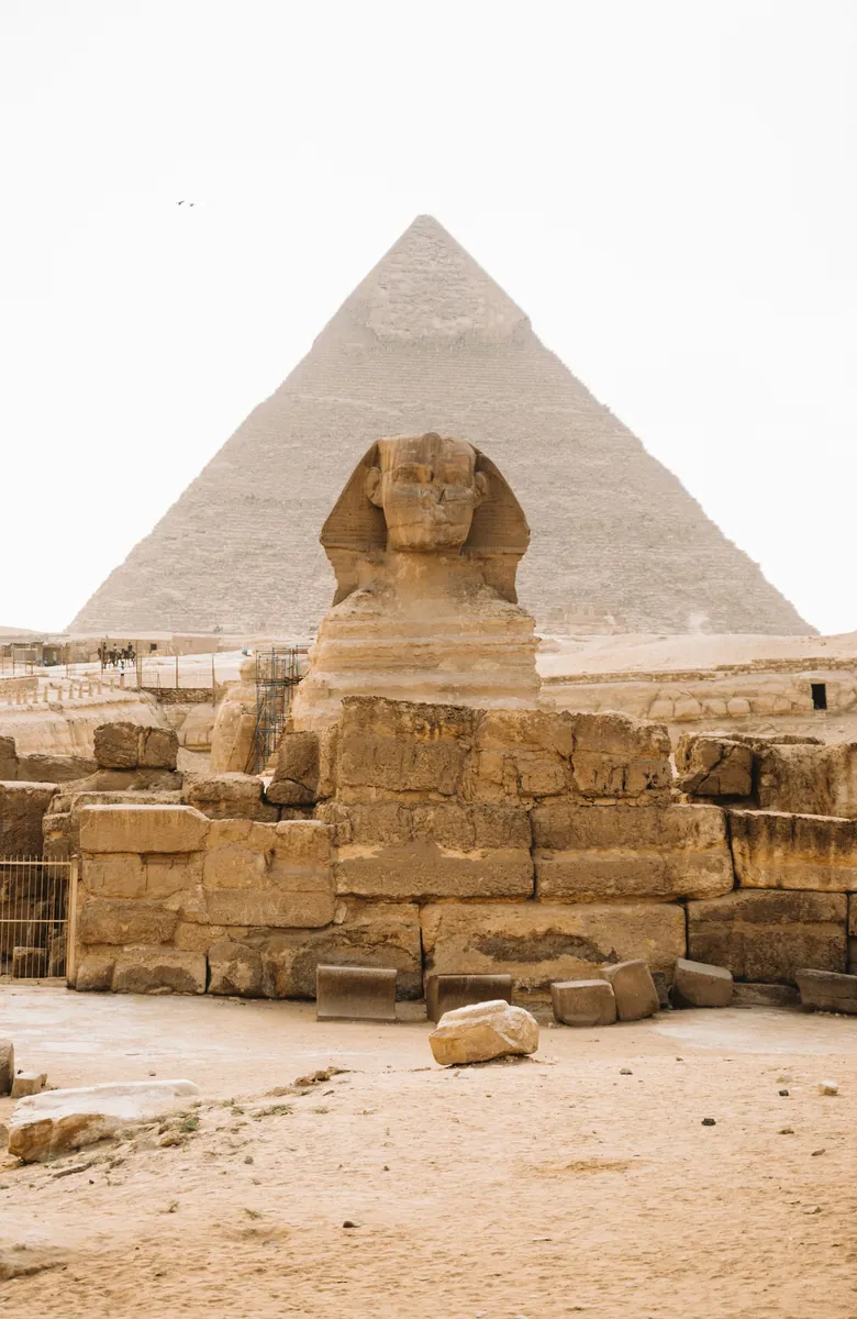 Große Sphinx von Gizeh vor der Chephren-Pyramide, davor verwitterte Steinblöcke unter hellem, diesigem Himmel
