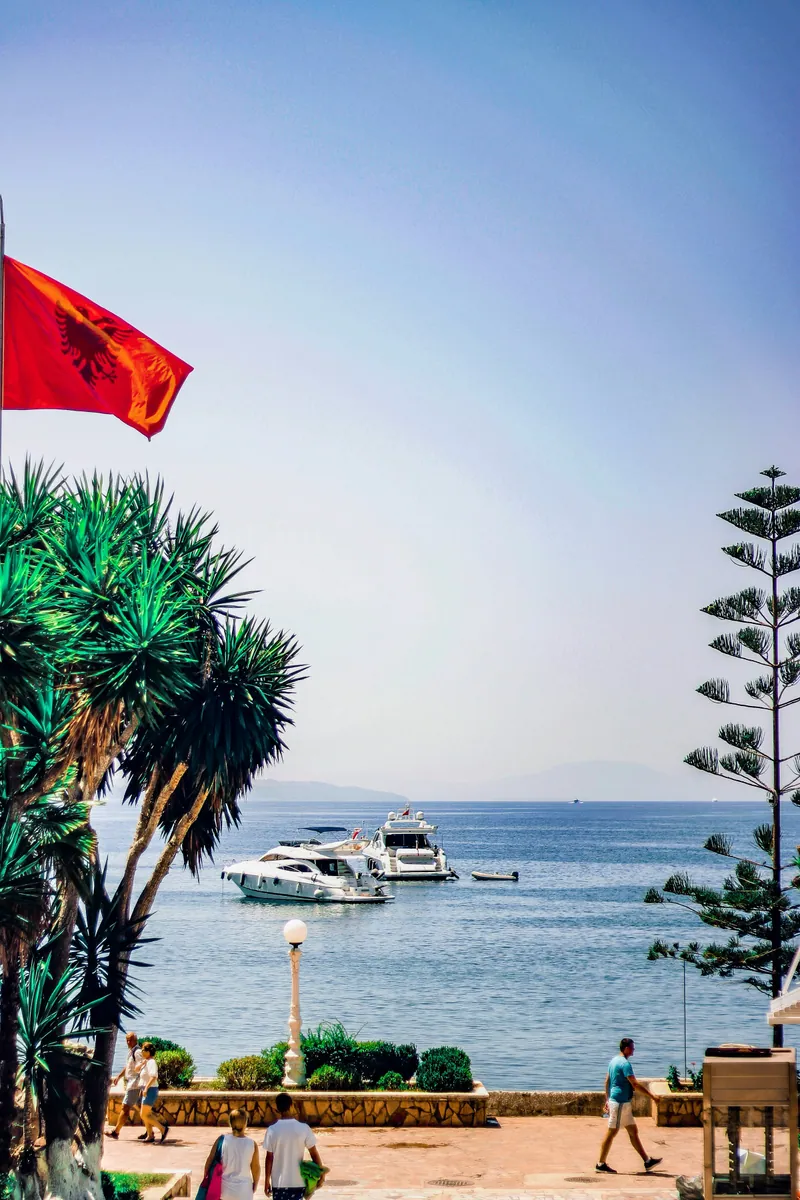 Albanische Flagge und Palmen an einer sonnigen Uferpromenade, in der Bucht ankern weiße Motoryachten vor fernen Bergen