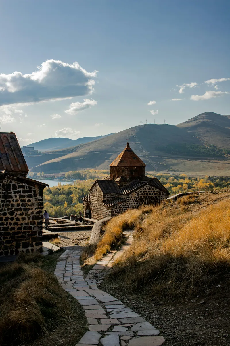 Gepflasterter Weg zu einer alten Steinkirche über einem See, umgeben von herbstlichen Bäumen und kahlen Hügeln