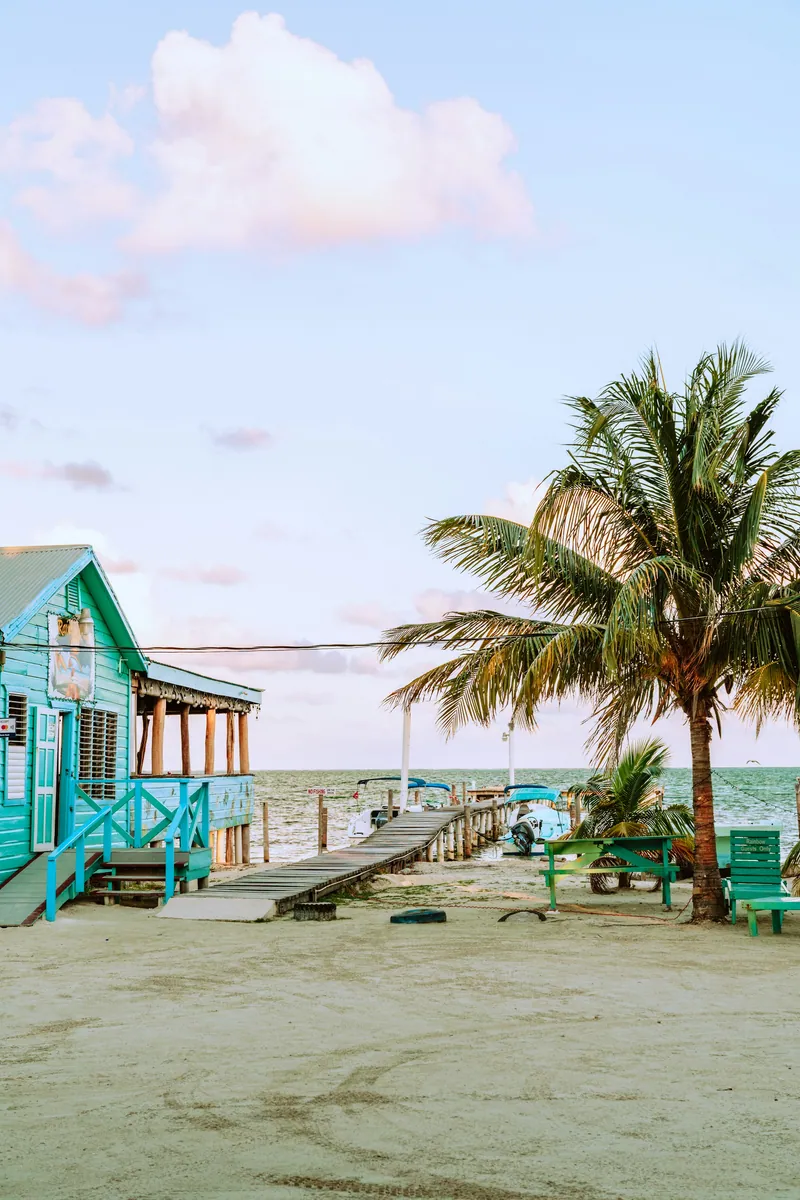 Türkisfarbenes Holzhaus mit Veranda am Sandstrand, daneben ein Holzsteg ins Meer, eine Palme und grüne Picknicktische
