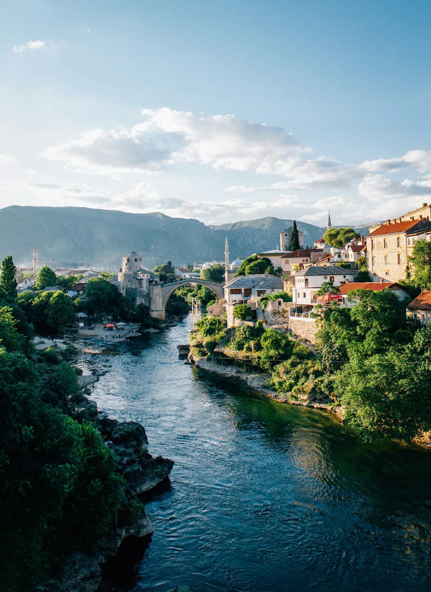 Alte Brücke von Mostar über die grüne Neretva, umgeben von Steinhäusern, Minaretten und bewaldeten Bergen
