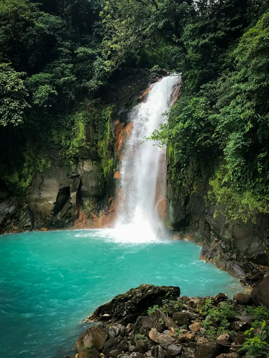 Wasserfall stürzt über dunkle, rostfarbene Felsen in ein leuchtend türkisblaues Becken inmitten von dichtem Regenwald