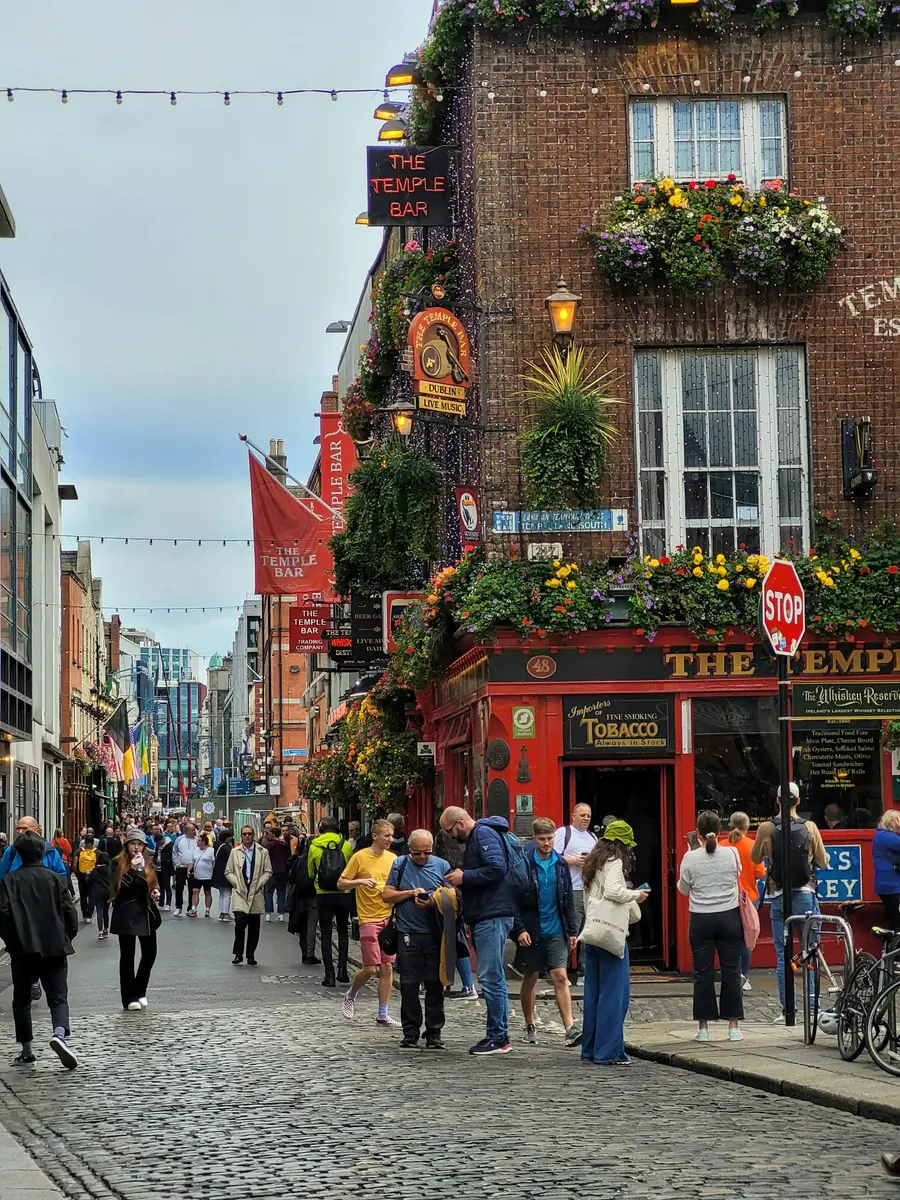 Pub The Temple Bar in Dublin mit Blumenschmuck an der roten Fassade, davor Passanten auf einer Kopfsteinpflastergasse