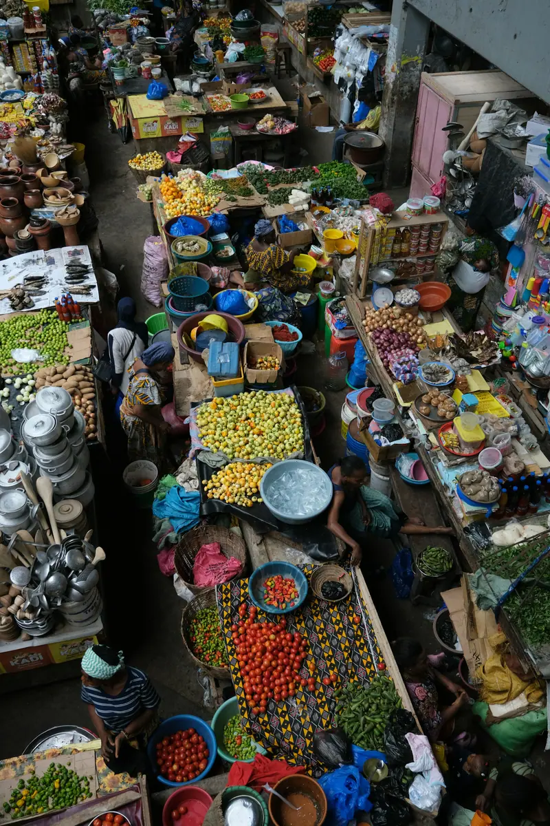 Blick von oben auf einen belebten Markt mit Tomaten, Chilischoten, gelbem Gemüse und Kochgeschirr an den Ständen