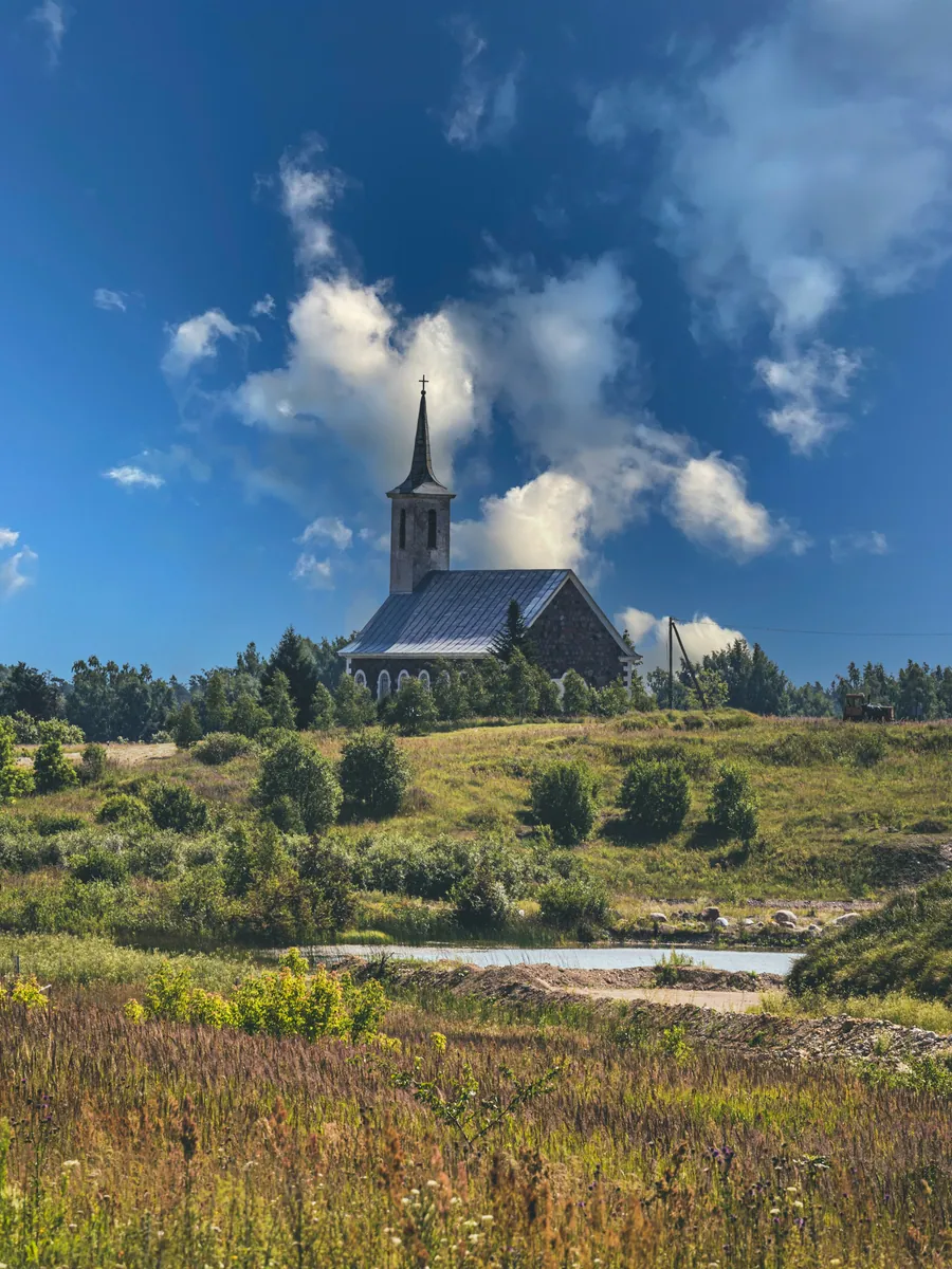 Steinkirche mit spitzem Turm auf einer Anhöhe über blühender Wiese und einem kleinen Teich unter Quellwolken