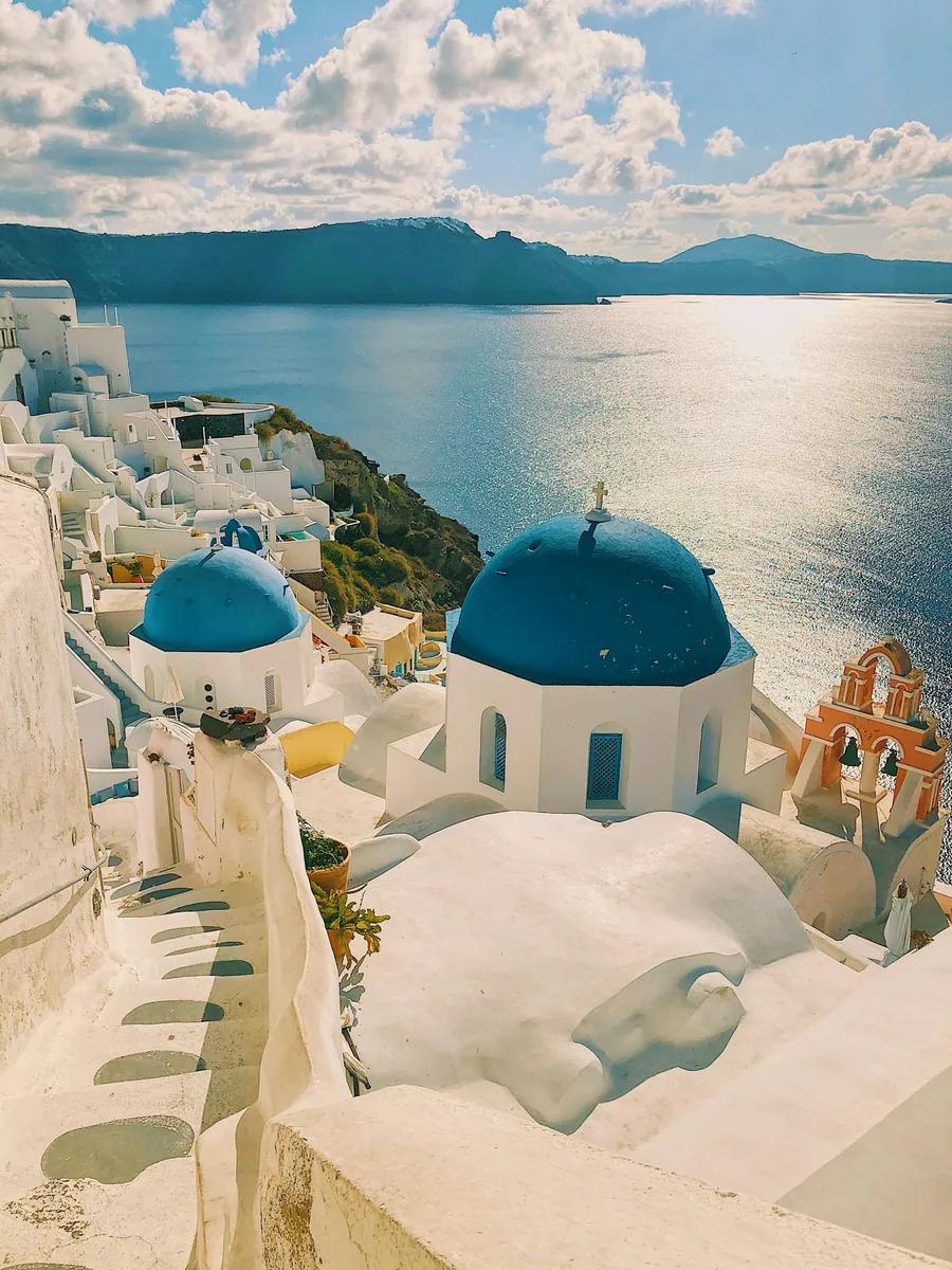 Blaue Kirchenkuppeln und weiße Häuser am Rand der Caldera von Santorini, dahinter das sonnenbeschienene Meer