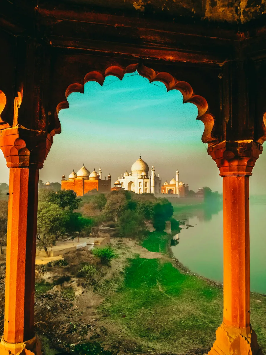 Das Taj Mahal und ein roter Sandsteinbau am Ufer der Yamuna, gerahmt von einem geschwungenen Zierbogen