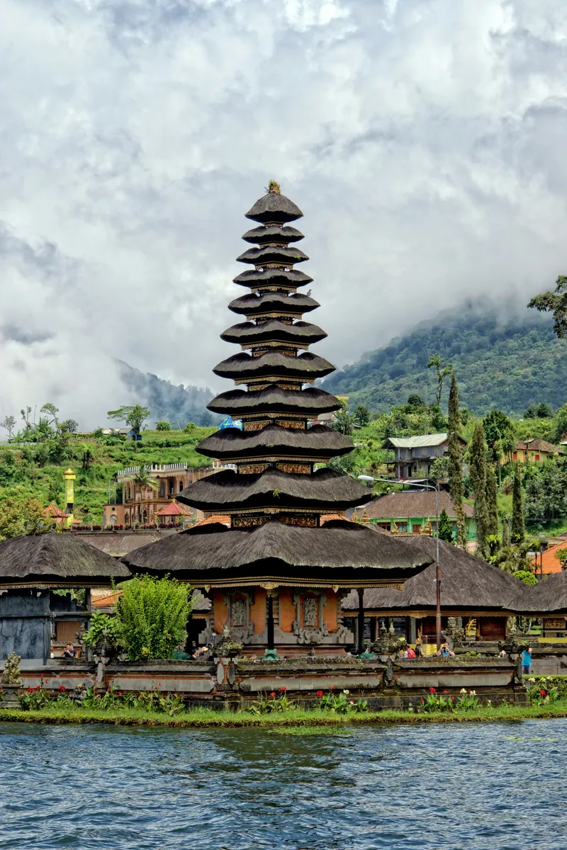 Mehrstufiger Meru-Turm des Tempels Pura Ulun Danu Bratan am Seeufer vor wolkenverhangenen grünen Bergen