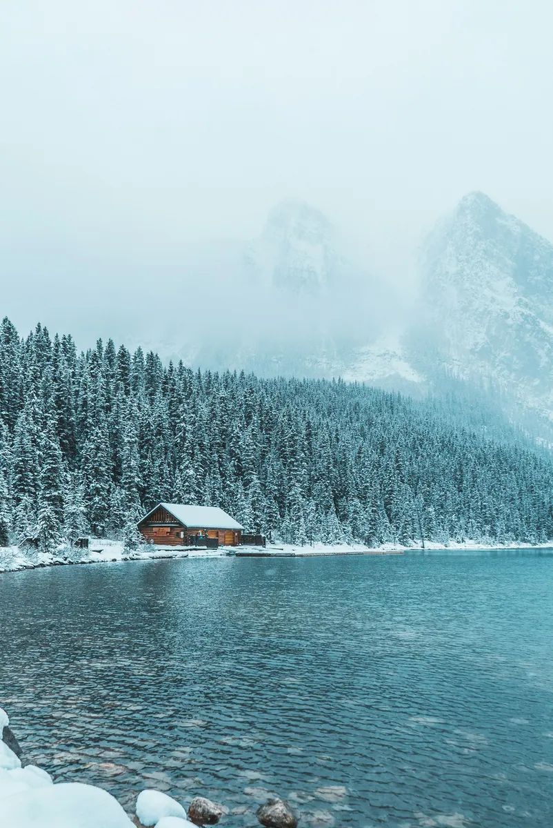 Holzbootshaus am Ufer eines Bergsees, dahinter verschneiter Nadelwald und in Nebel gehüllte Gipfel