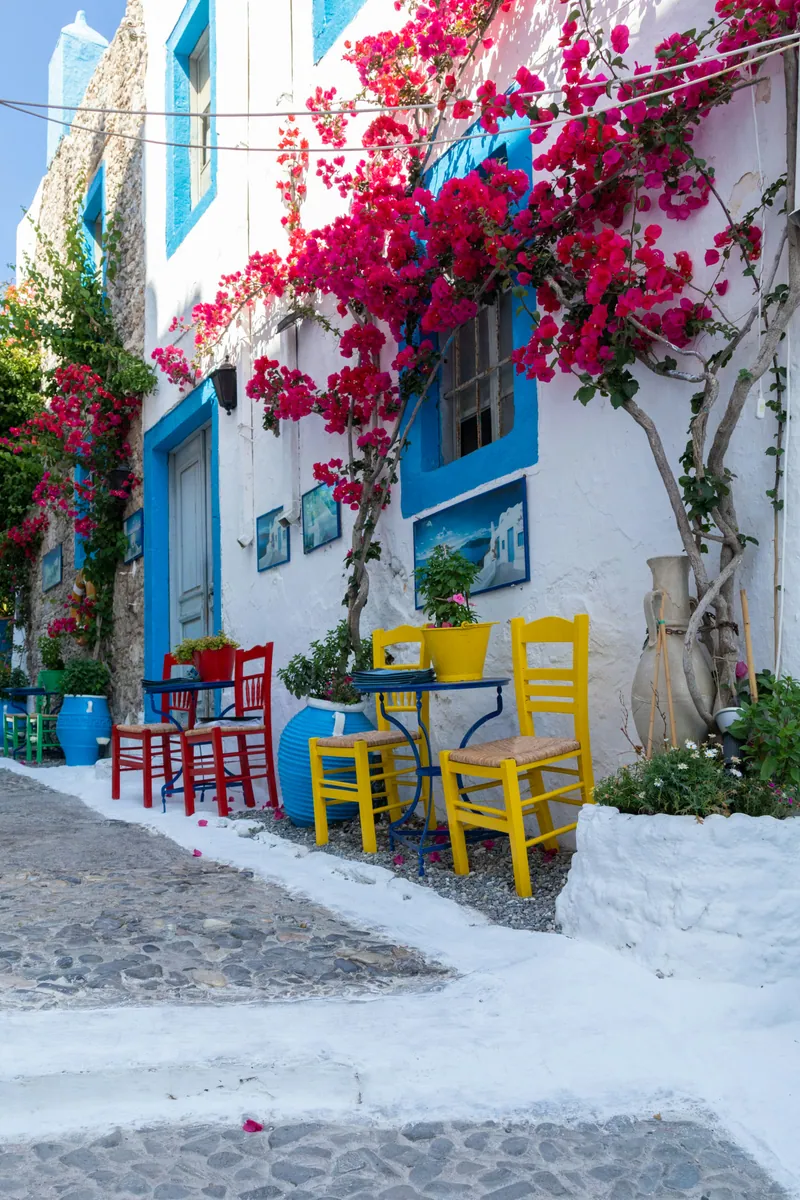 Weiß getünchte Gasse mit blauen Fensterrahmen, pinker Bougainvillea und bunten Stühlen an kleinen Tischen