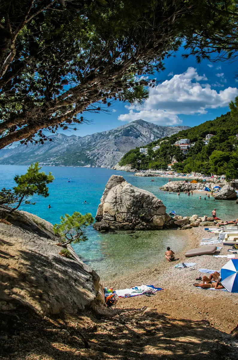 Kiesstrand mit großem Felsblock im türkisfarbenen Meer, Pinien und Badegäste, dahinter ein kahles Küstengebirge