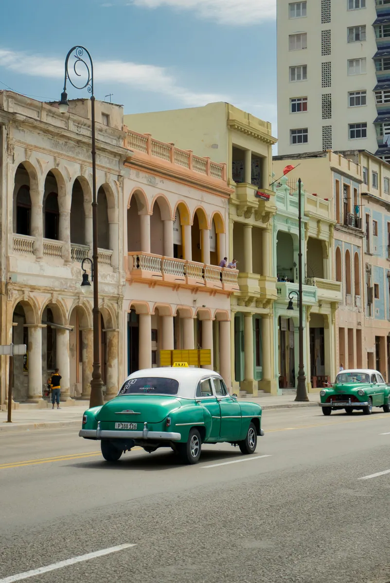 Grüne Oldtimer aus den 1950er-Jahren, einer davon ein Taxi, vor pastellfarbenen Häusern mit Arkaden