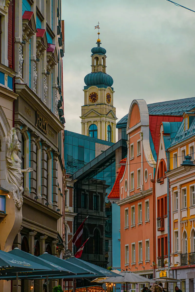 Bunte Altstadtfassaden mit Skulptur und Sonnenschirmen, dahinter ein Turm mit grüner Haube und Uhr