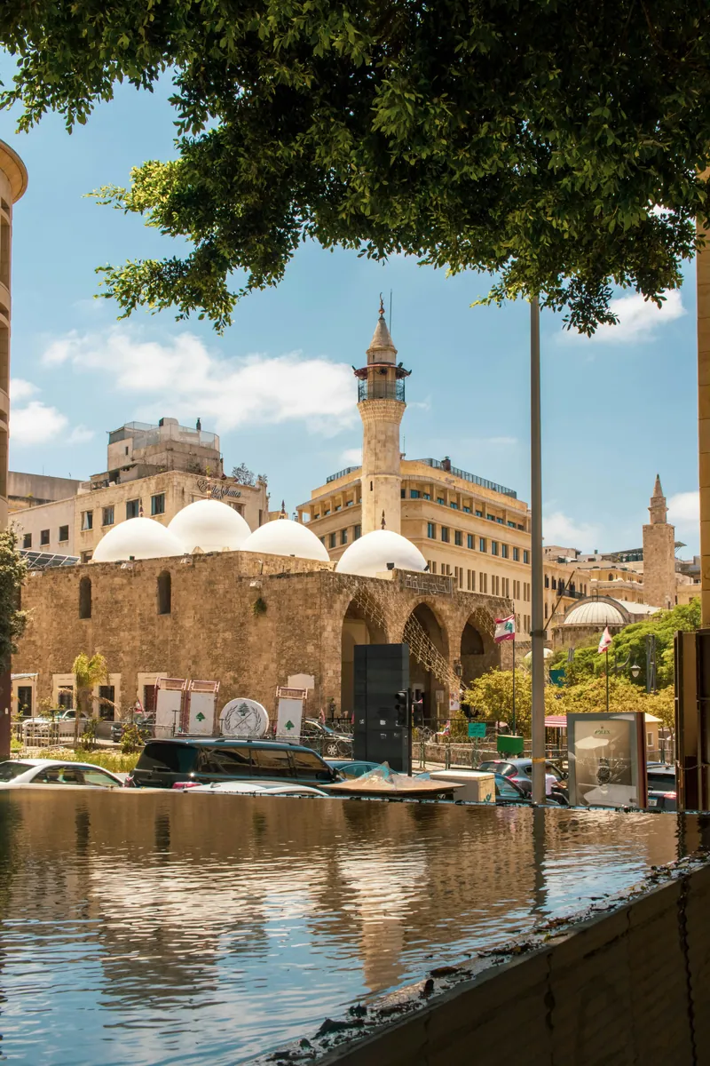 Steinbau mit weißen Kuppeln und Minarett, davor Autos und libanesische Flaggen, im Vordergrund spiegelndes Wasser