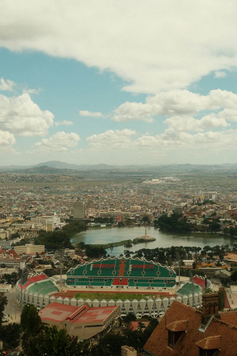 Blick über Antananarivo mit dem Anosy-See und seinem Denkmal, davor ein grün-rotes Stadion, Berge am Horizont