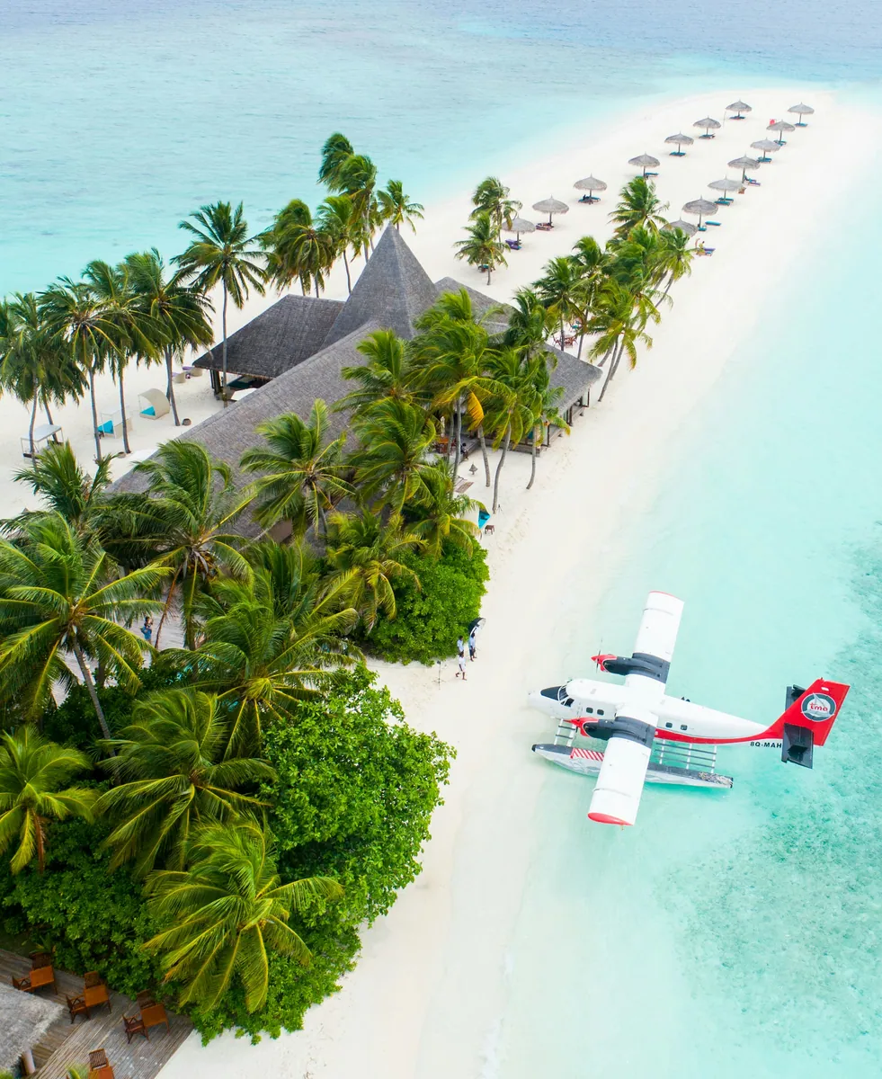 Wasserflugzeug im türkisfarbenen Wasser vor einer Insel mit weißem Sandstrand, Palmen und Strohdachbauten