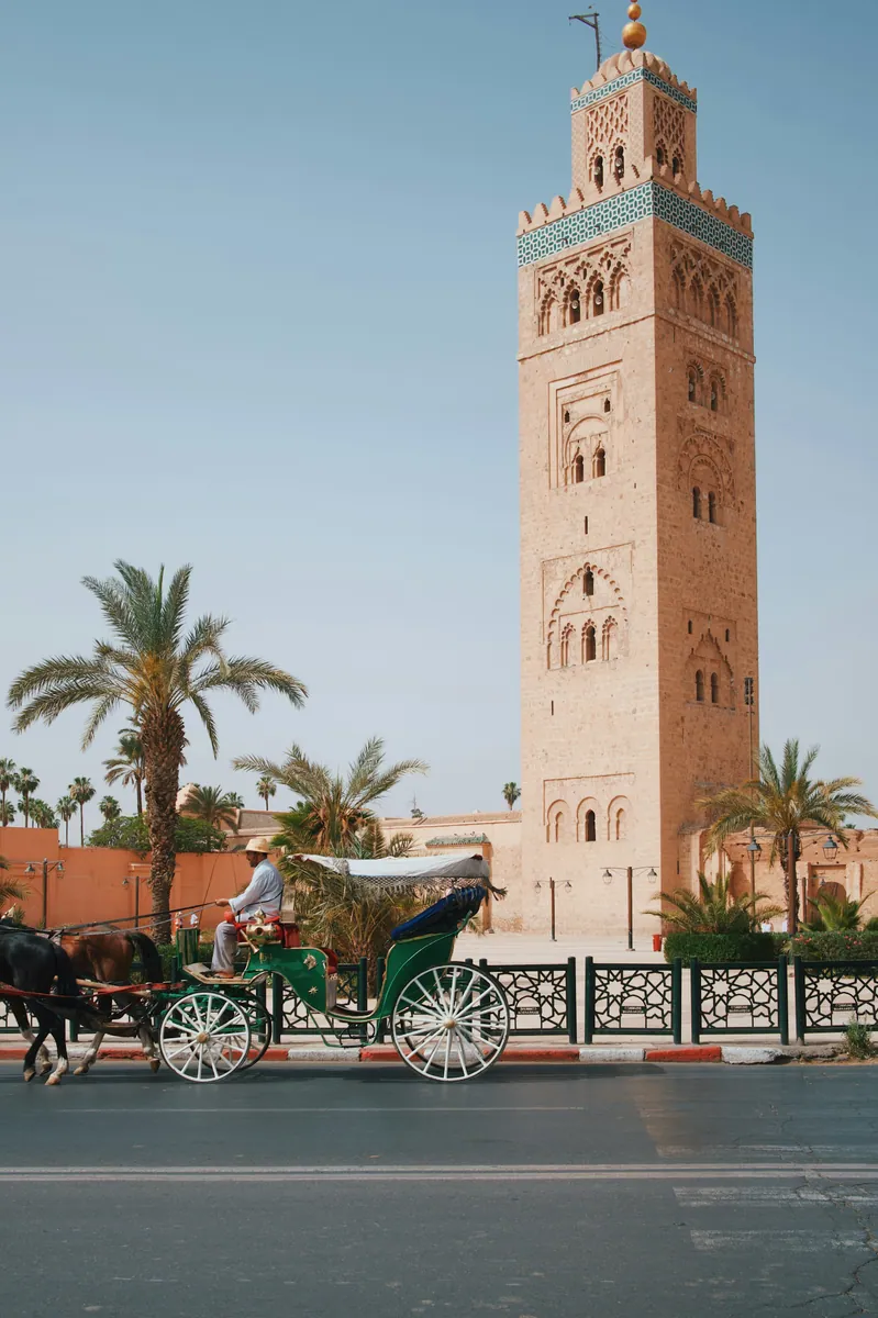 Minarett der Koutoubia-Moschee in Marrakesch zwischen Palmen, davor eine grüne Pferdekutsche auf der Straße