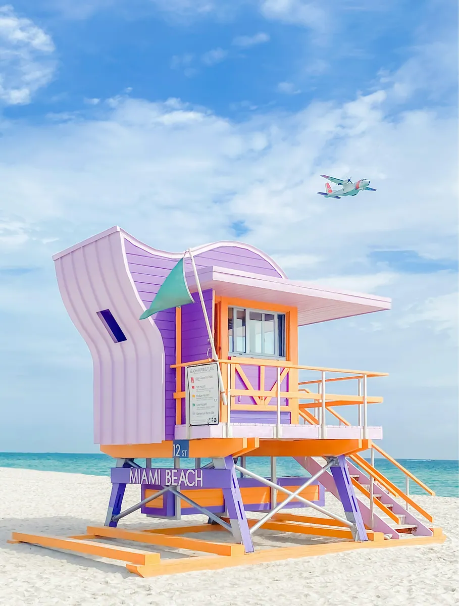 Lila-orangefarbener Rettungsschwimmerturm mit der Aufschrift Miami Beach am weißen Sandstrand vor türkisem Meer