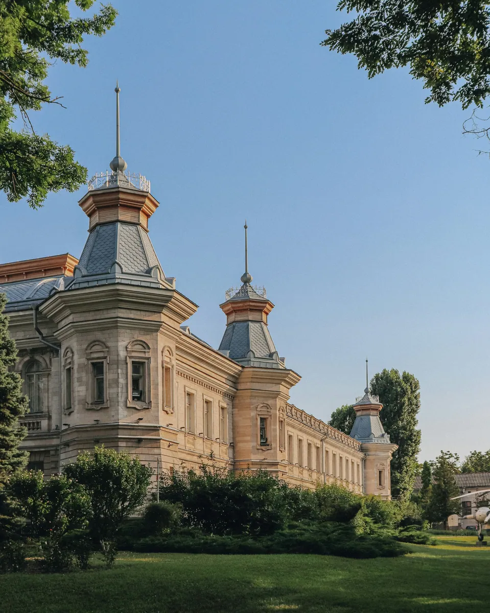 Historisches Gebäude mit Türmchen unter grauen Spitzhauben hinter Bäumen und einer gepflegten Rasenfläche