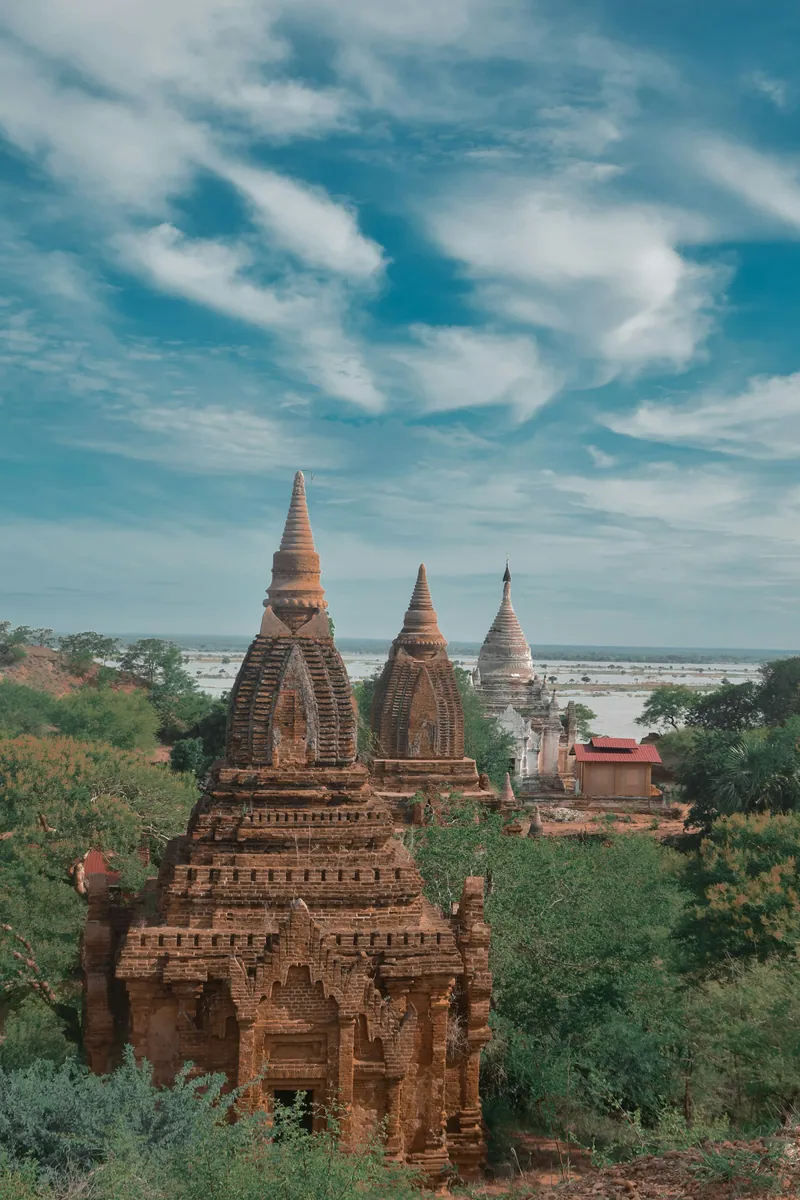 Alte Backsteinpagoden und eine weiße Stupa zwischen Bäumen, im Hintergrund eine weite Wasserfläche