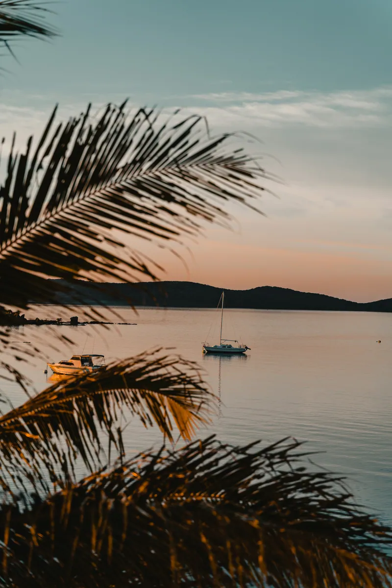 Segelboot und Motorboot in einer ruhigen Bucht in der Abenddämmerung, im Vordergrund Palmwedel