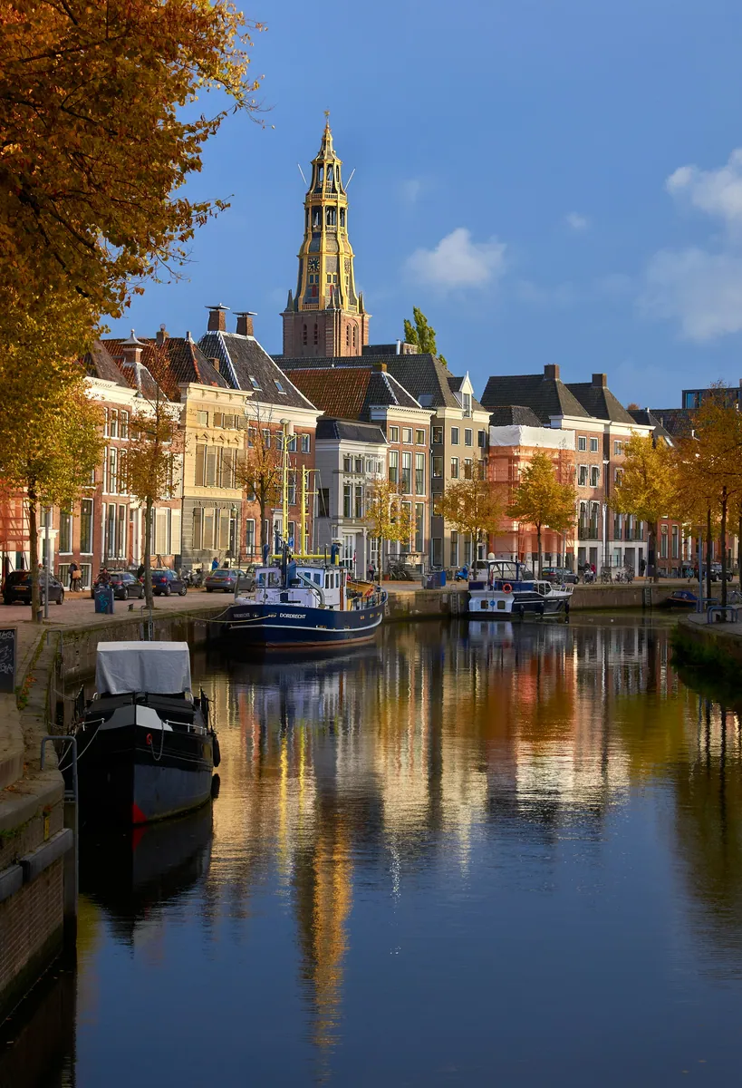 Gracht mit Booten und Backsteinhäusern im Herbst, dahinter ein Kirchturm, alles spiegelt sich im Wasser