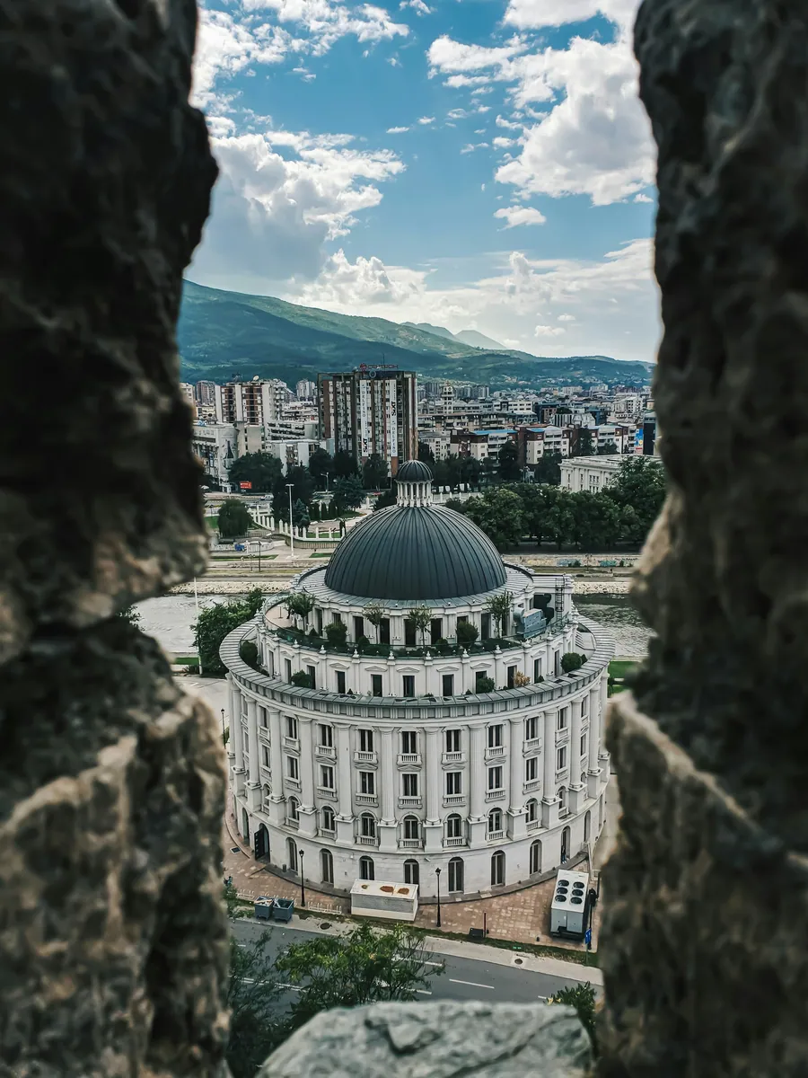 Blick durch eine Lücke in einer alten Steinmauer auf ein rundes weißes Kuppelgebäude, dahinter Stadt und grüne Berge.