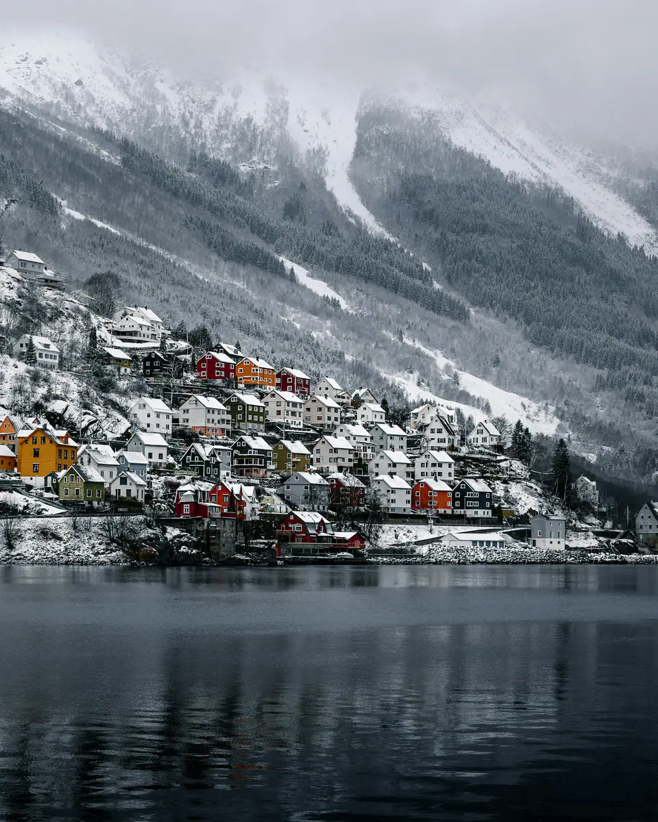 Bunte Holzhäuser auf einem verschneiten Hang am Ufer eines Fjords, darüber nebelverhangene Berge im Winter.