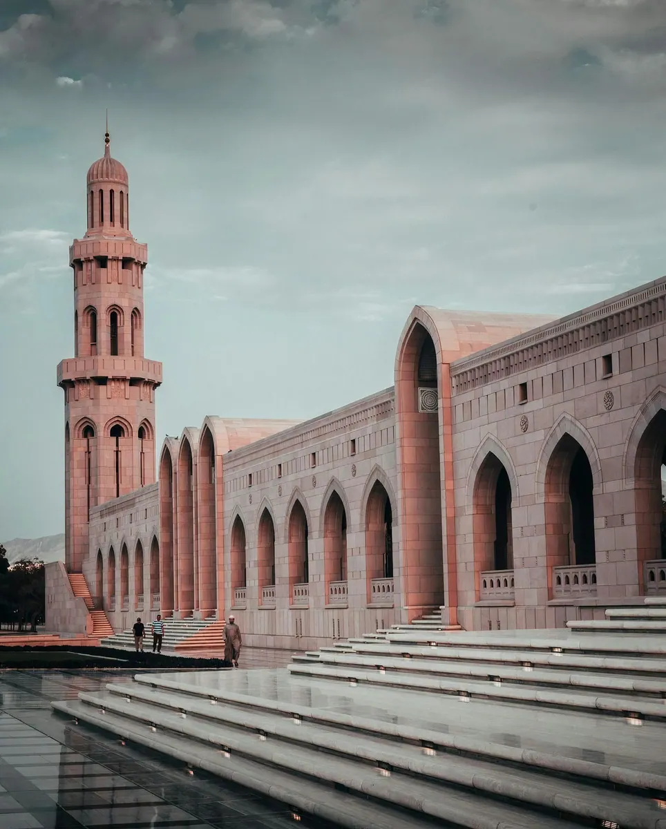 Rosafarbenes Minarett und lange Spitzbogen-Arkaden einer großen Moschee, im Vordergrund helle Marmorstufen.