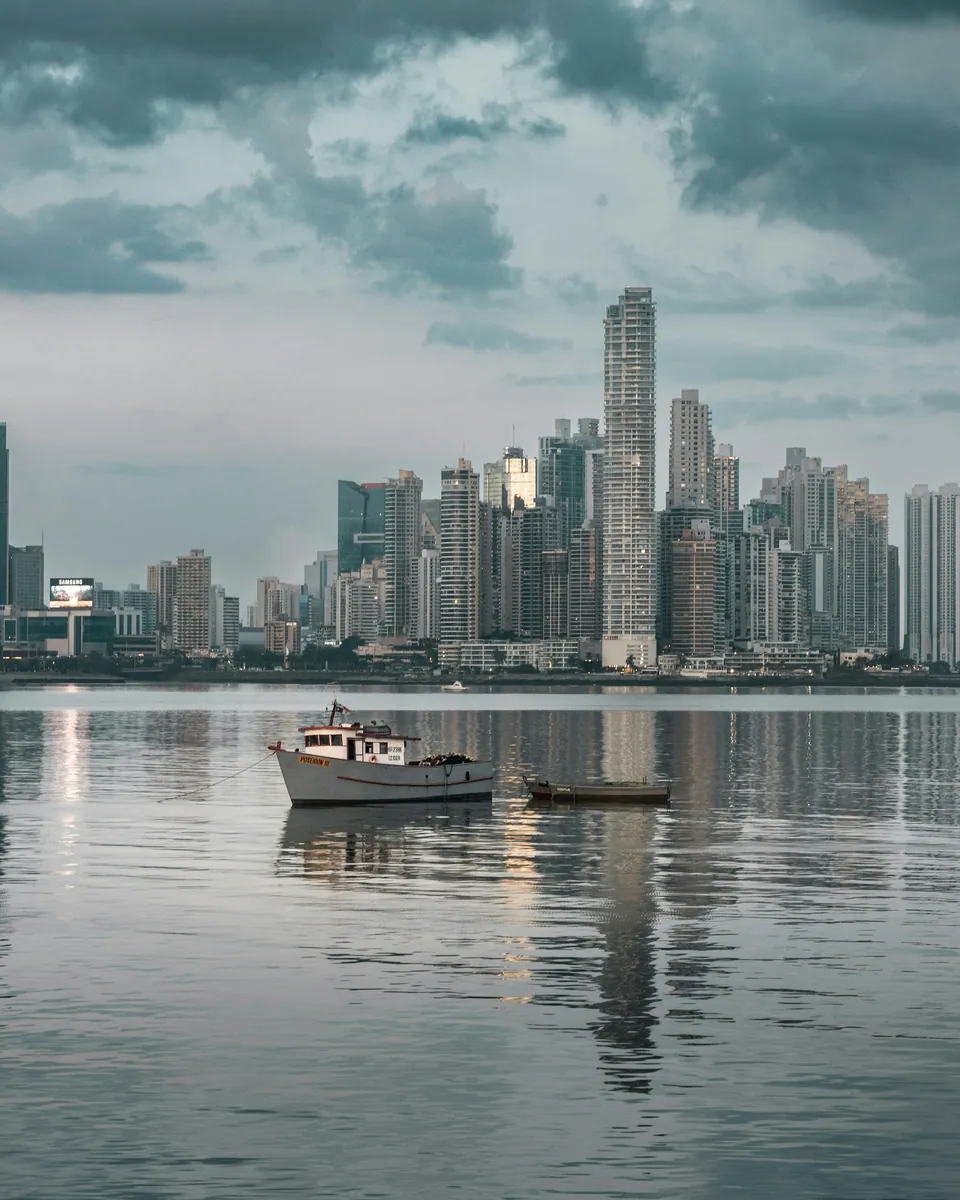 Ein Fischerboot und ein kleines Holzboot auf ruhigem Wasser vor einer Hochhaus-Skyline unter dunklen Wolken.