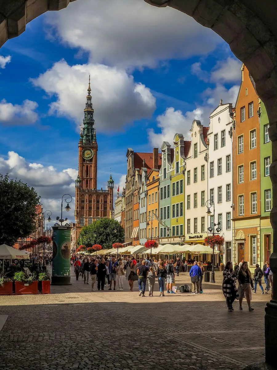 Blick durch einen Torbogen auf eine belebte Straße mit bunten Giebelhäusern und dem Turm des Rechtstädtischen Rathauses in Danzig.
