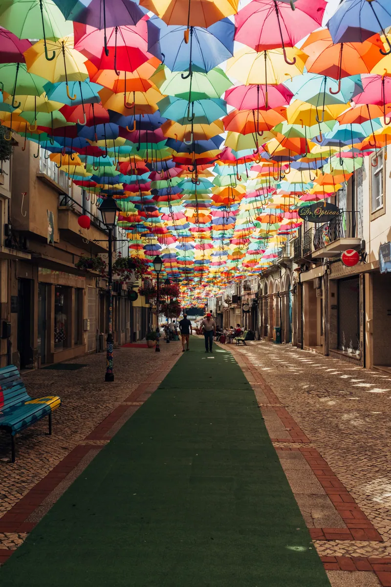 Hunderte bunte Regenschirme hängen über einer gepflasterten Fußgängerstraße mit grünem Teppichstreifen.
