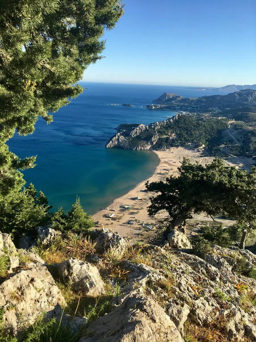 Blick von einem felsigen Hügel mit Nadelbäumen auf eine Sandbucht mit Sonnenschirmen und tiefblaues Meer