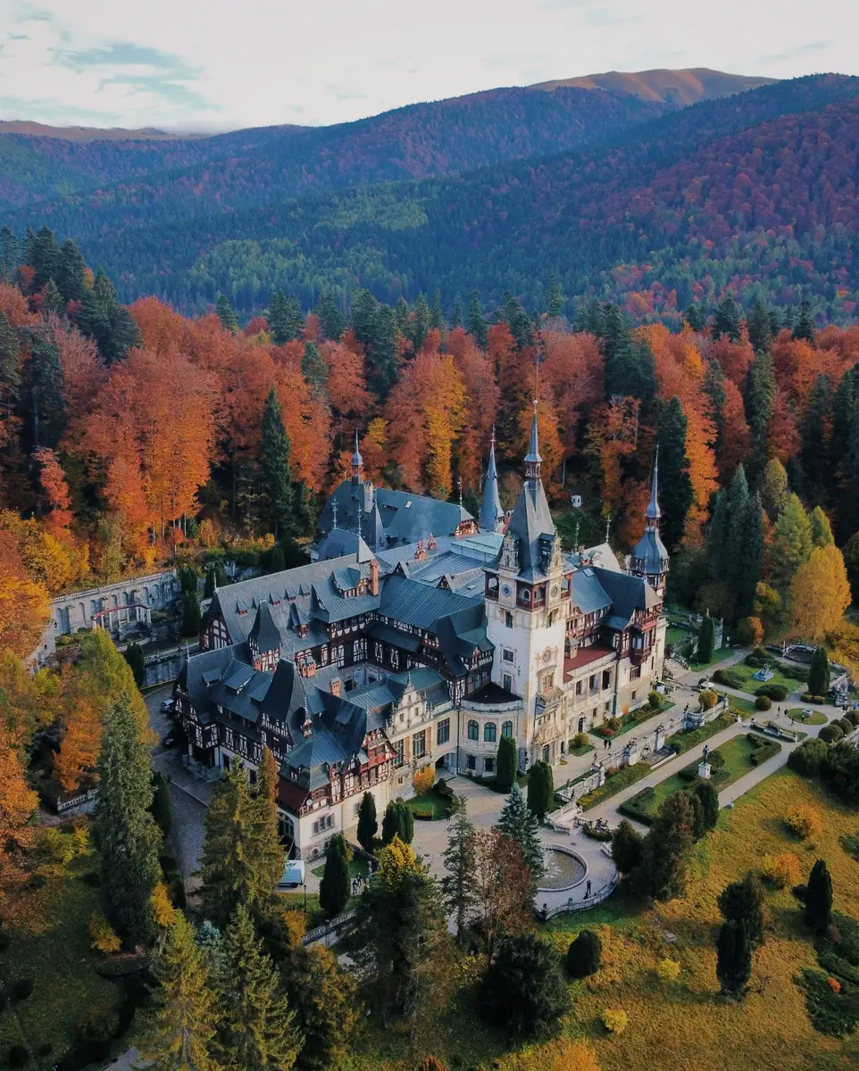 Luftaufnahme von Schloss Peleș mit Türmen und Fachwerkfassaden, umgeben von Herbstwald in Rot und Orange.