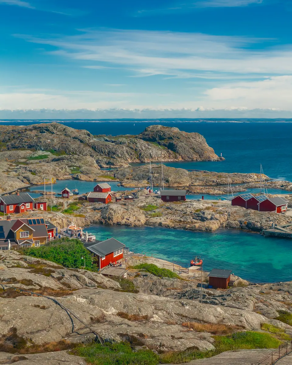 Rote Holzhäuser zwischen glatten Granitfelsen an einer türkisfarbenen Bucht, dahinter Schären und offenes Meer.
