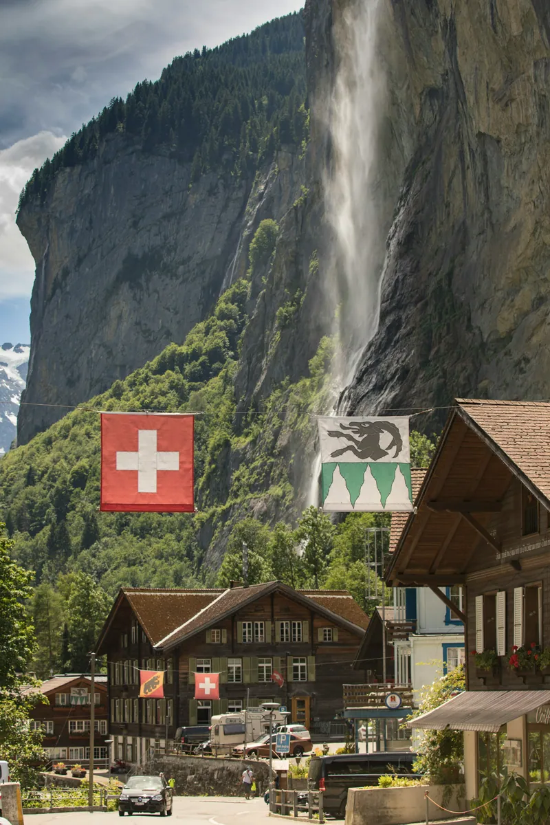 Der Staubbachfall stürzt eine Felswand oberhalb von Lauterbrunnen hinab, davor Holzchalets und Fahnen über der Dorfstraße.