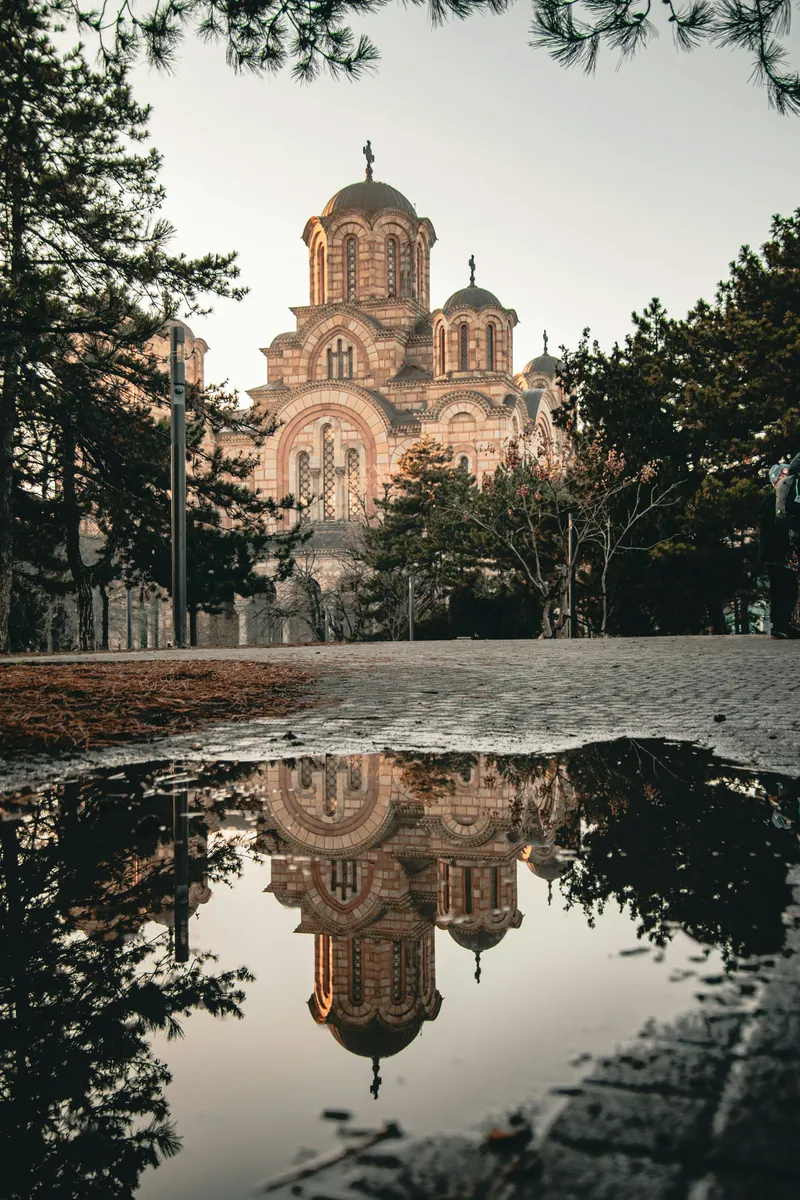 Backsteinkirche im byzantinischen Stil mit Kuppeln, gespiegelt in einer Pfütze auf einem Parkweg zwischen Kiefern.