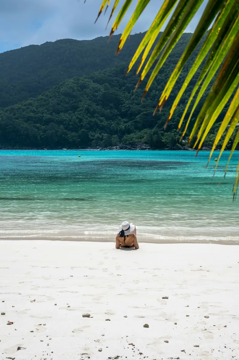 Eine Person mit weißem Sonnenhut sitzt am weißen Sandstrand vor türkisfarbenem Wasser und bewaldeten Hügeln.