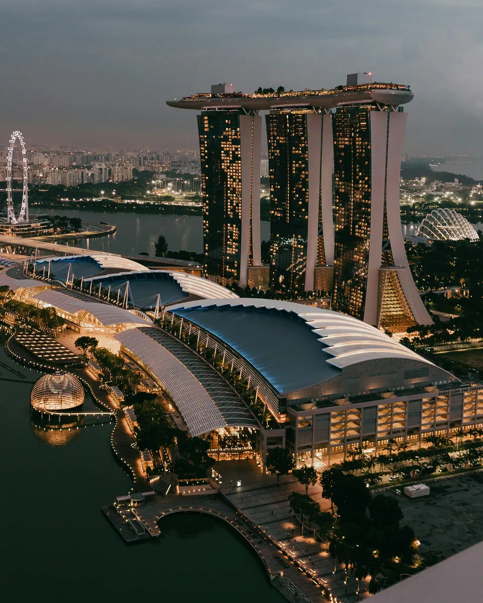 Marina Bay Sands mit Dachpark über dem beleuchteten Ufer der Marina Bay in Singapur in der Abenddämmerung, links ein Riesenrad
