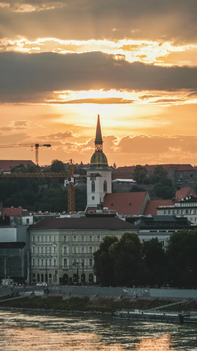 Kirchturm mit grüner Kuppel und spitzem Helm über Dächern am Flussufer, daneben Baukräne im Sonnenuntergang.