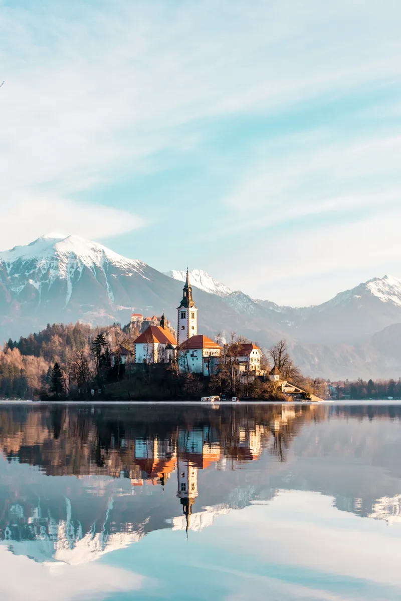 Die Inselkirche im Bleder See spiegelt sich im stillen Wasser vor schneebedeckten Gipfeln der Julischen Alpen.