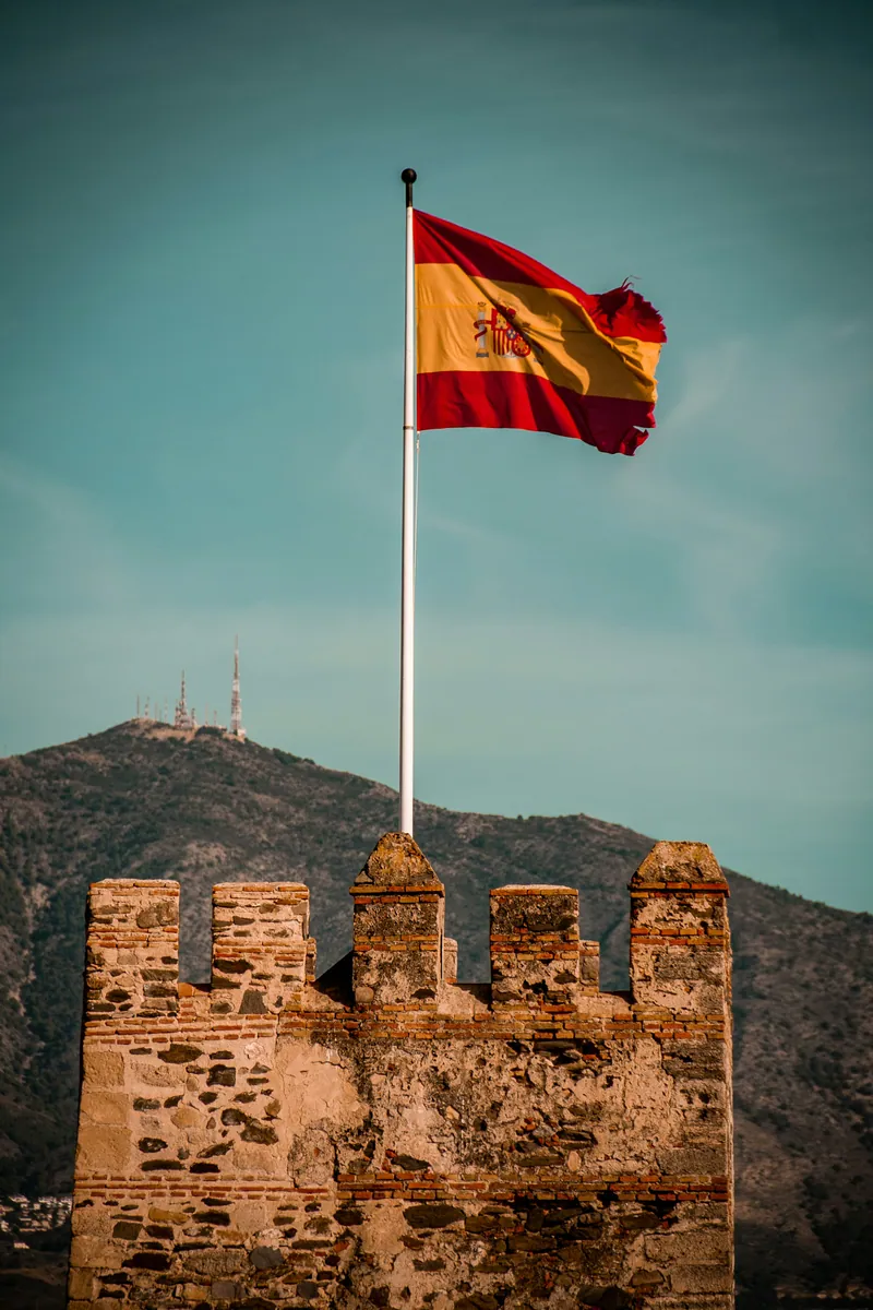 Die spanische Flagge weht über den Zinnen eines alten Steinturms, dahinter ein Berg mit Sendemasten.