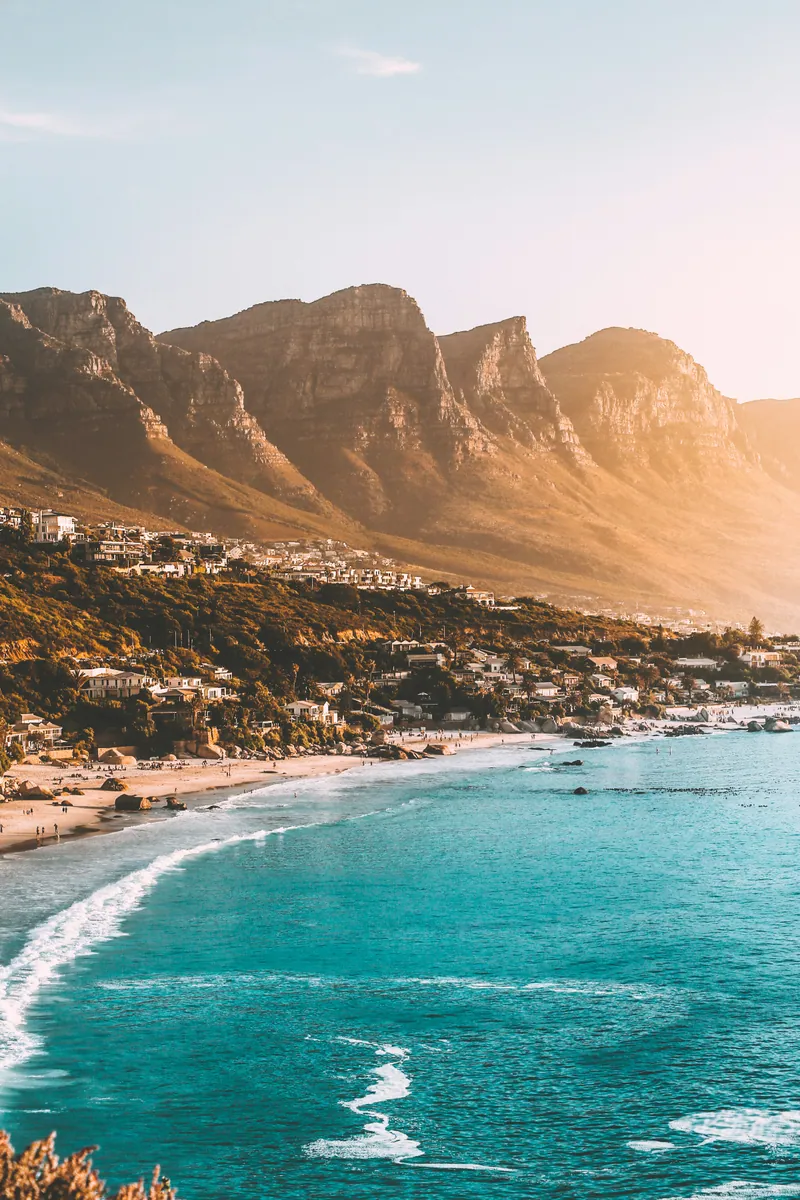 Abendlicht über einer Bucht mit türkisfarbenem Wasser und Sandstrand, darüber Villen am Hang und eine schroffe Bergkette.