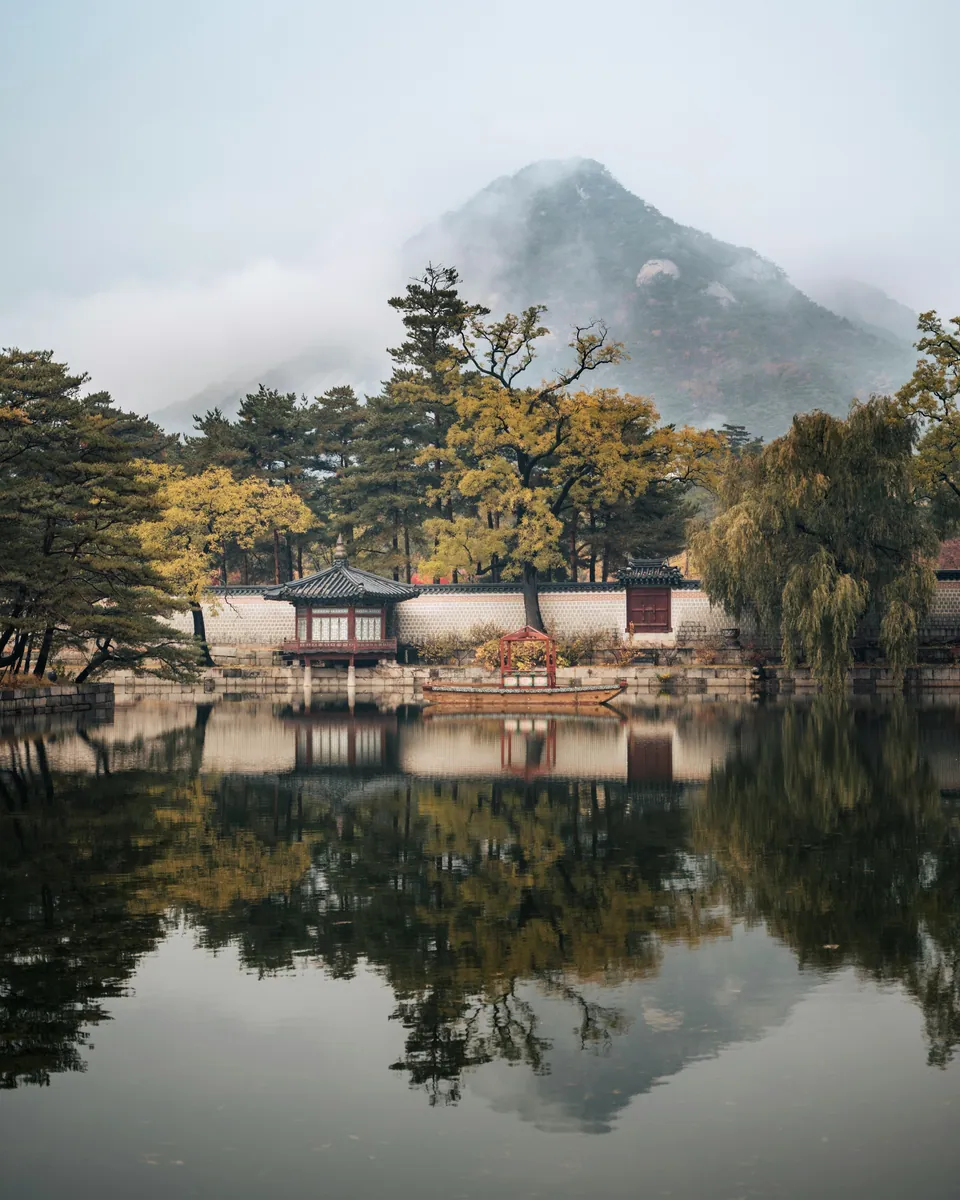 Pavillon mit geschwungenem Ziegeldach an einer Palastmauer spiegelt sich in einem Teich, dahinter ein Berg im Nebel.