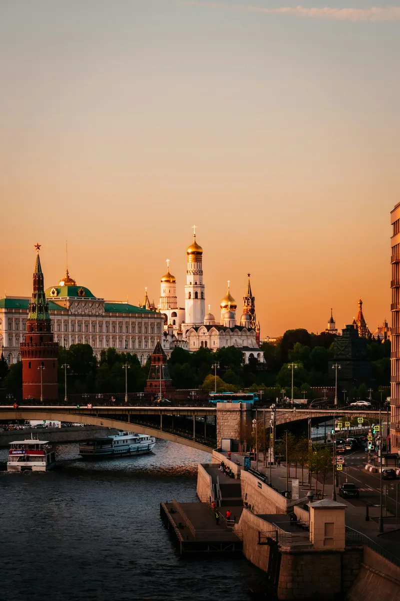 Moskauer Kreml mit Großem Kremlpalast und Glockenturm Iwan der Große bei Sonnenuntergang, davor die Moskwa mit Brücke und Schiffen