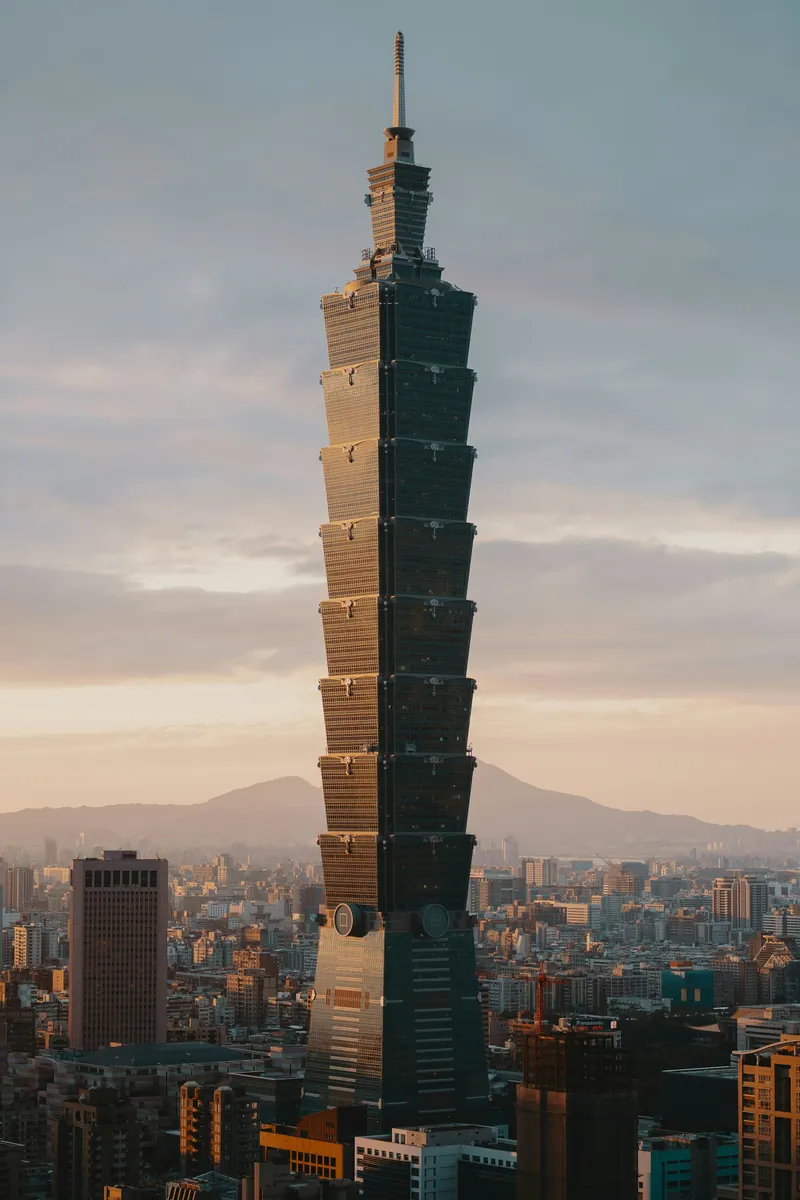 Der Wolkenkratzer Taipei 101 ragt im warmen Licht der tief stehenden Sonne über die Stadt, dahinter Berge.