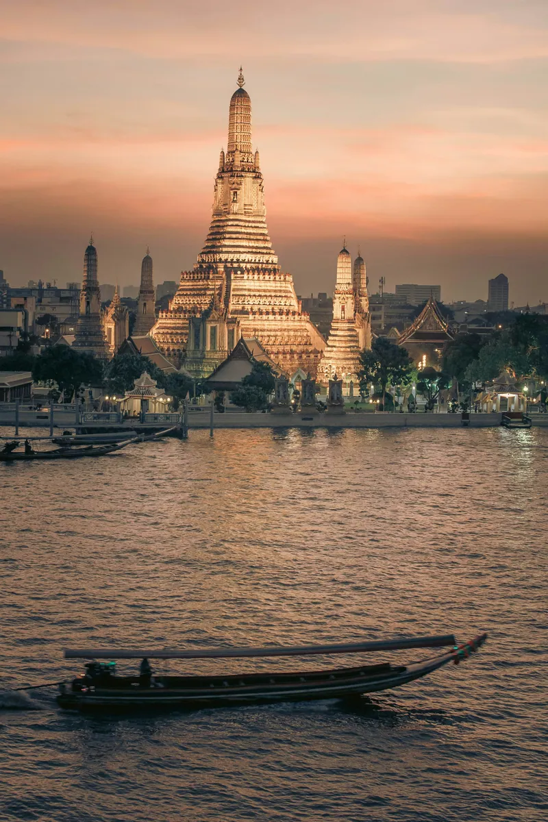 Tempel Wat Arun in Bangkok in der Abenddämmerung, davor ein Longtail-Boot auf dem Fluss Chao Phraya
