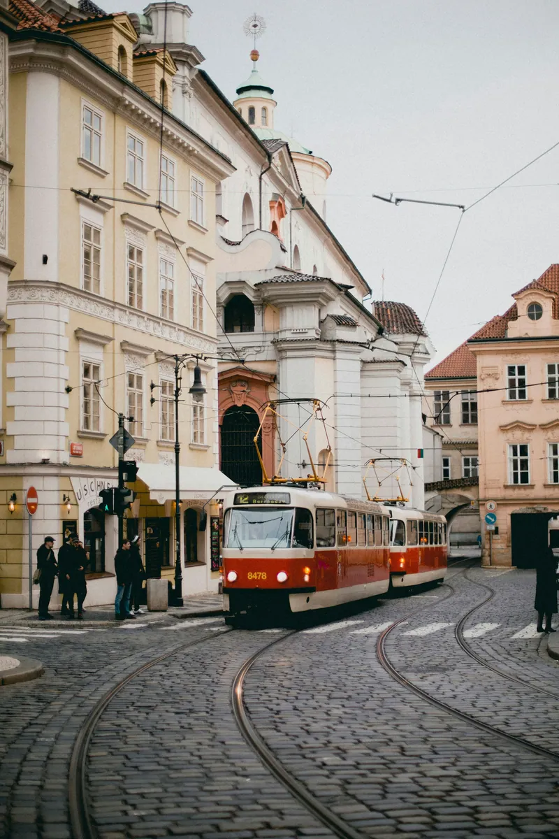 Rot-weiße Straßenbahn auf einer gepflasterten Kreuzung in Prag vor barocken Bürgerhäusern und einer Kirche