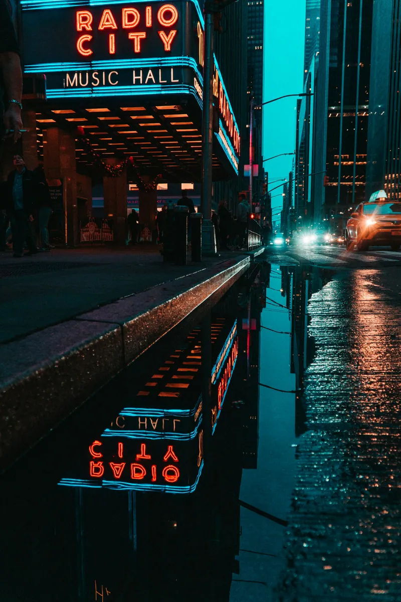Die Leuchtreklame der Radio City Music Hall in New York spiegelt sich nachts in einer Pfütze, daneben ein gelbes Taxi.