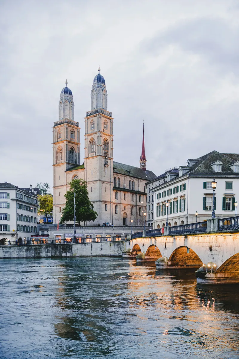 Grossmünster mit seinen zwei Türmen an der Limmat in Zürich, rechts eine beleuchtete Bogenbrücke an einem bewölkten Abend